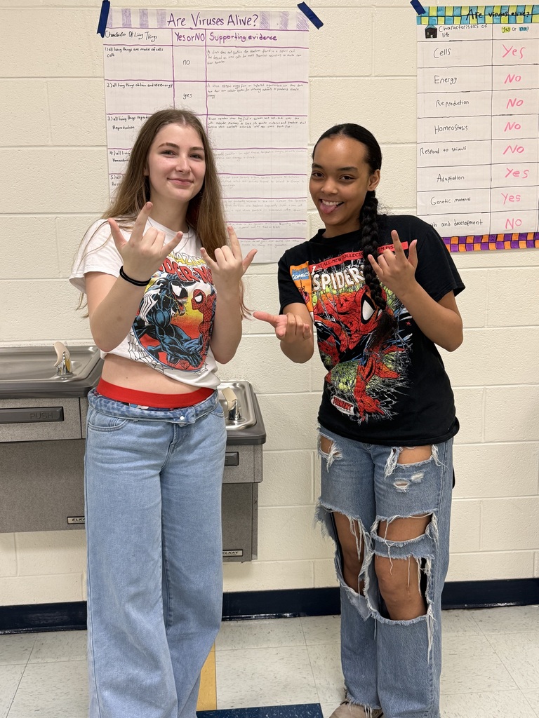 Two women standing in a classroom. One wears a white T-shirt, and the other a black T-shirt. Both have jeans, and one makes a peace sign.