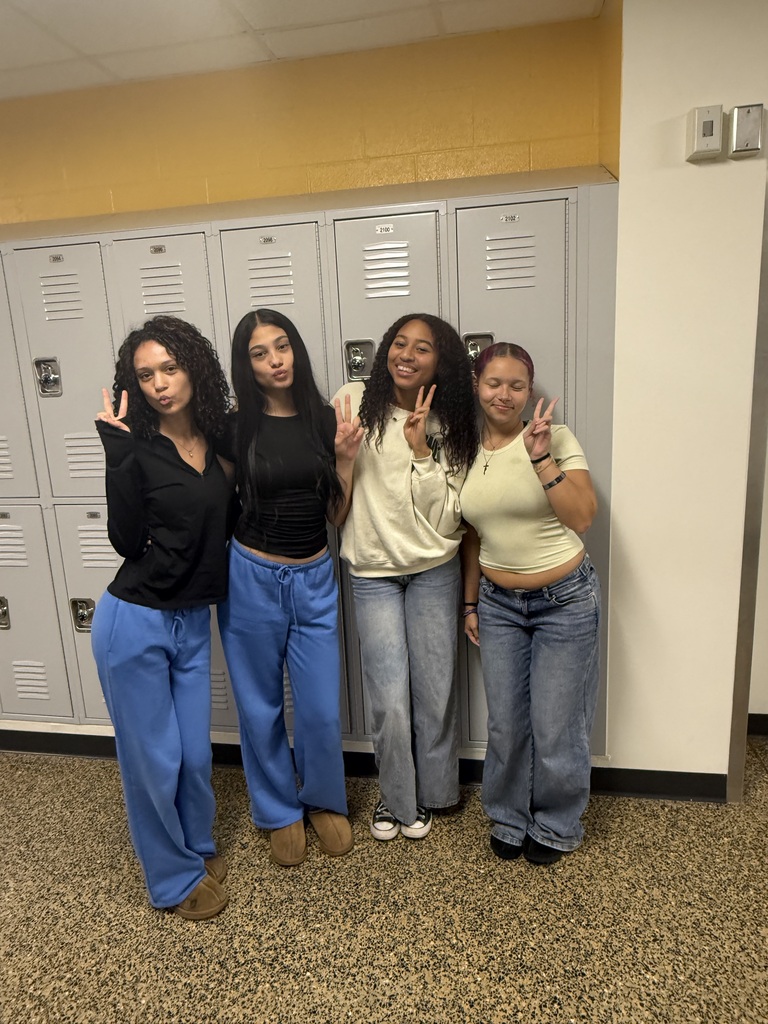 Four women standing together in front of lockers in a hallway, all making a peace sign with their hands.