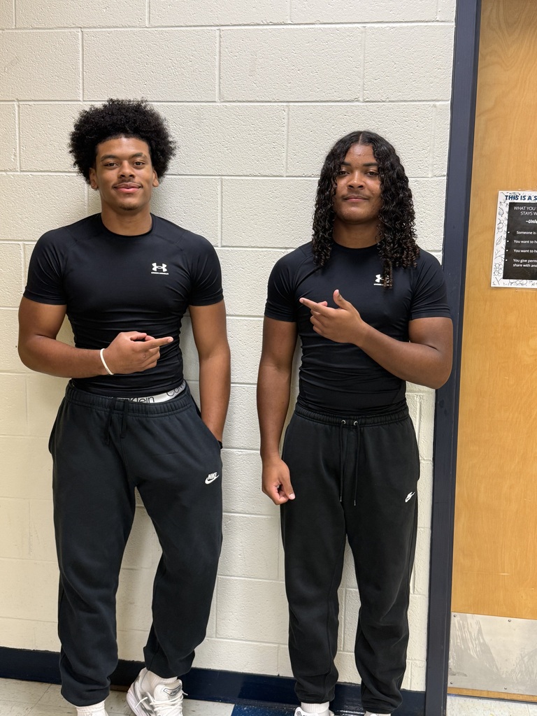 Two men stand next to each other in front of a white wall. Both are dressed in matching black t-shirts and pants.