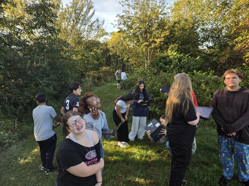 A group of people stand in a grassy area surrounded by trees and shrubs. Some individuals are wearing glasses.