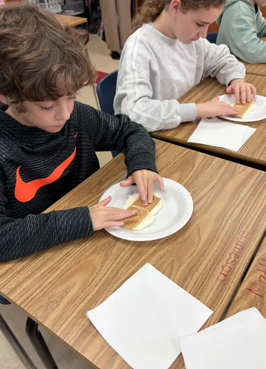 Two children seated at desks, one eating a sandwich. A blurred person is in the background.