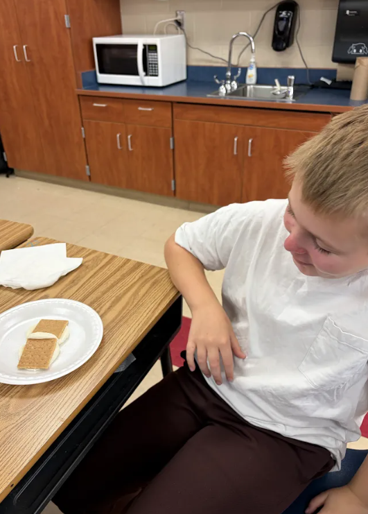 Boy sitting at a table, eating a sandwich, wearing a white shirt and brown pants.