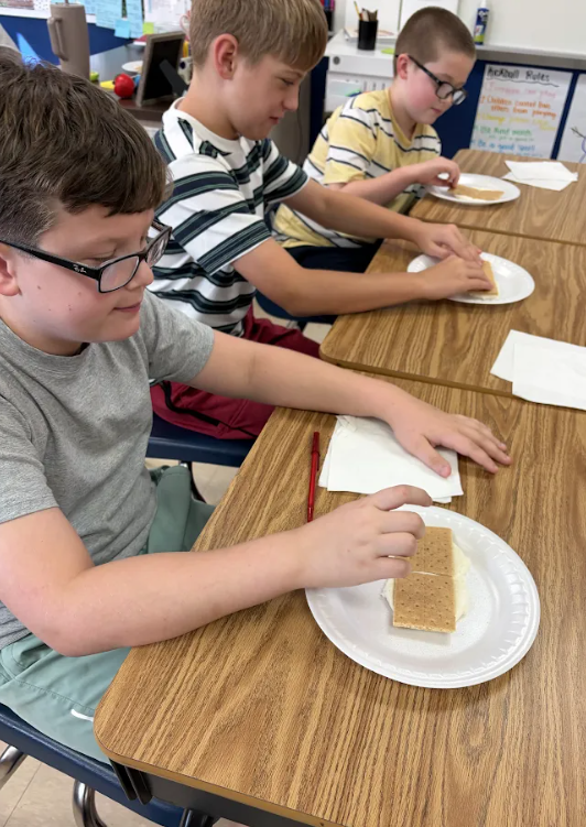 Three boys seated at a table eating s'mores. One holds a s'more on a plate.