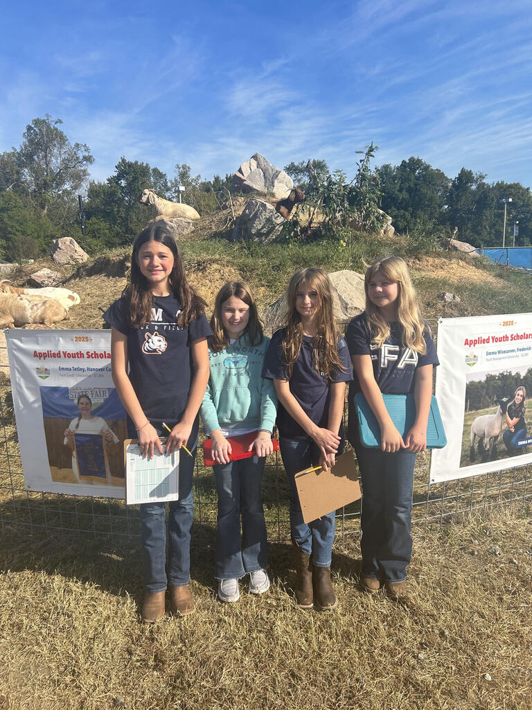 Four CMS FFA students pose together at the State Fair of Virginia in front of a livestock enclosure with goats standing on rocks in the background. The students are holding clipboards, papers, and pencils while participating in agricultural competitions.