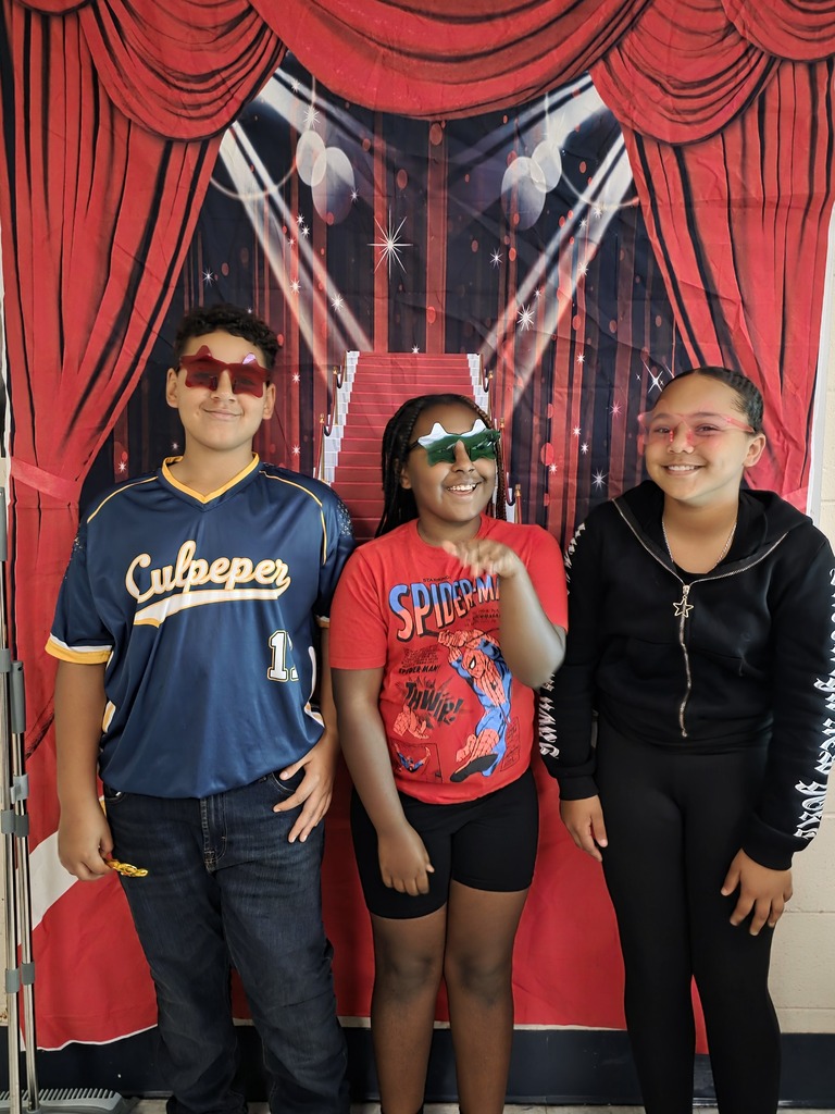 Three sixth-grade students wearing colorful star-shaped glasses smile and pose together in front of a red-carpet backdrop.