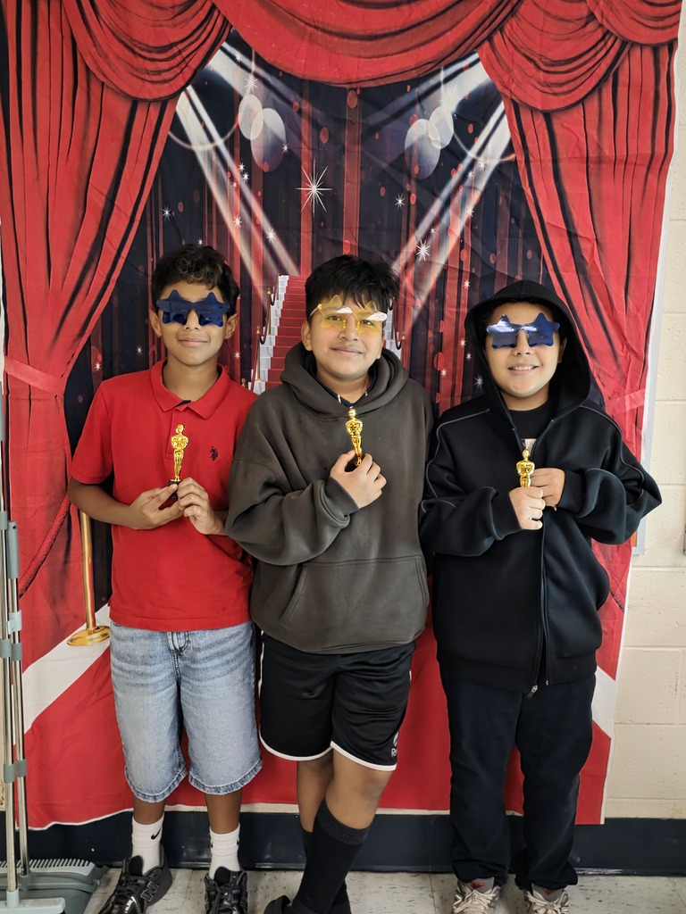 Three sixth-grade students wearing colorful star-shaped glasses pose with small gold trophies in front of a red-carpet backdrop.