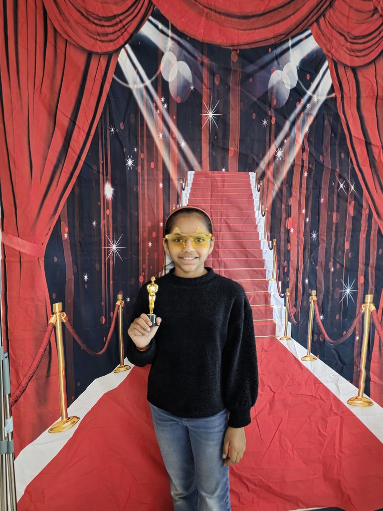 A sixth-grade student wearing gold star-shaped glasses smiles while holding a small gold trophy in front of a red-carpet backdrop.