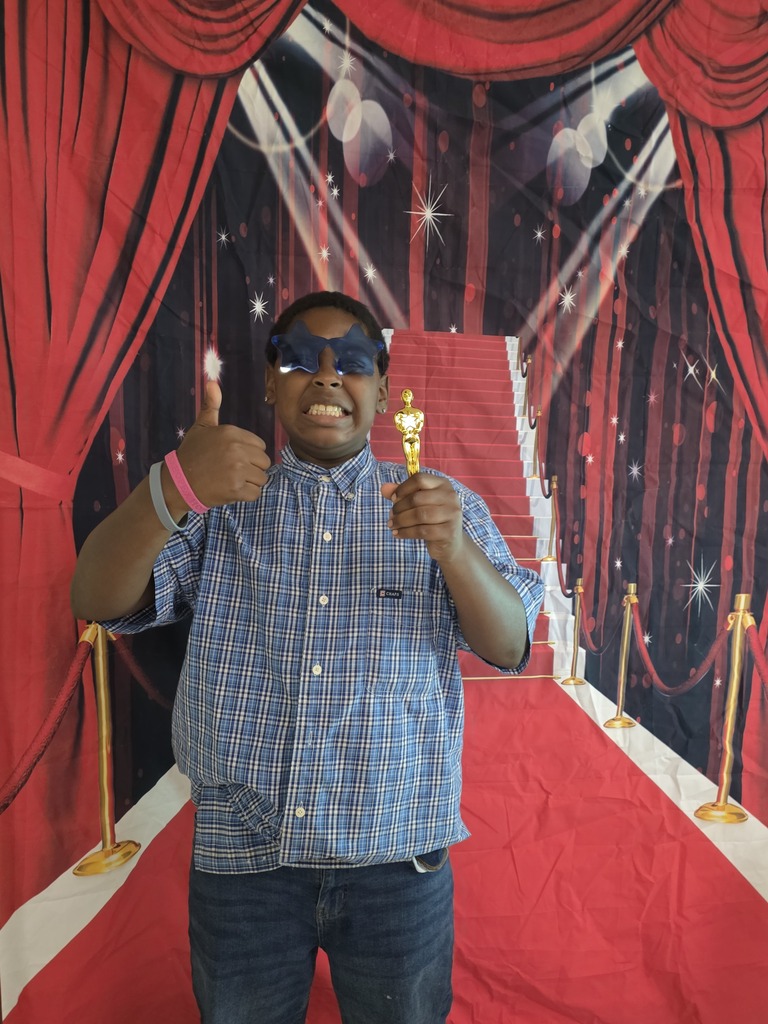 A sixth-grade student wearing blue star-shaped glasses smiles, gives a thumbs-up, and holds a small gold trophy while posing in front of a red-carpet backdrop.