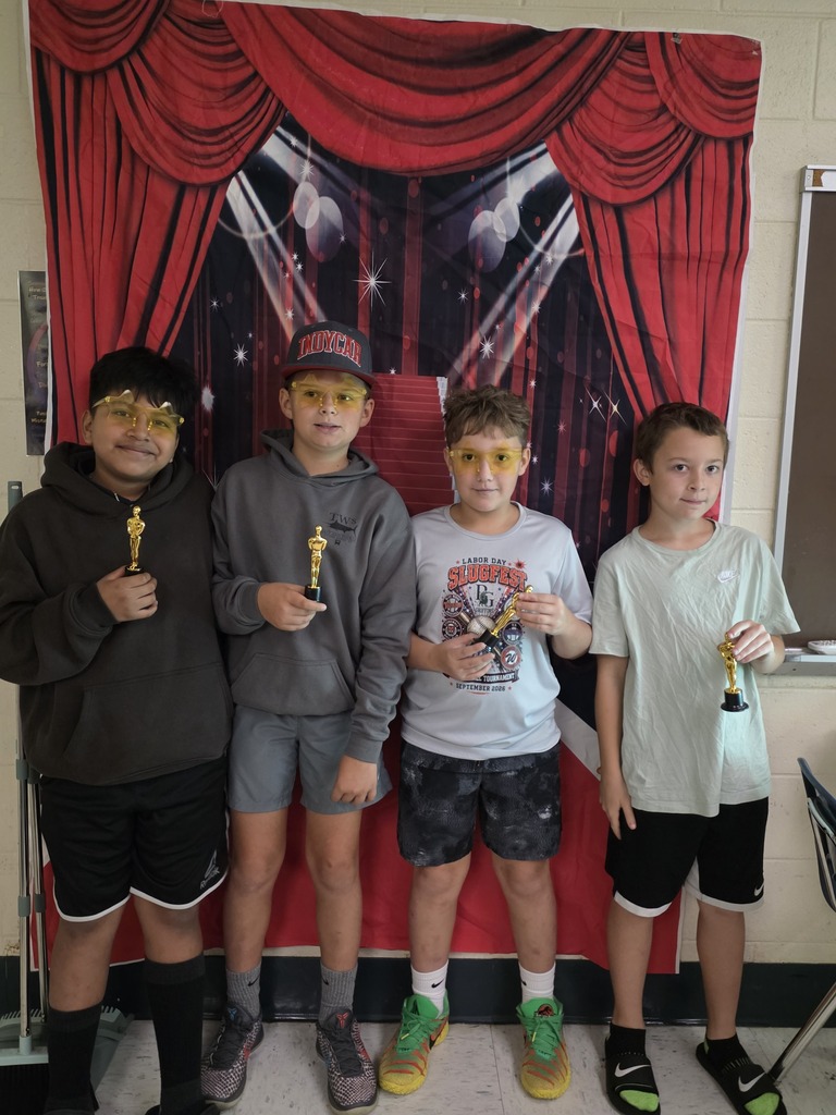Four sixth-grade students pose with small gold trophies in front of a red-carpet backdrop, with several wearing gold star-shaped glasses.
