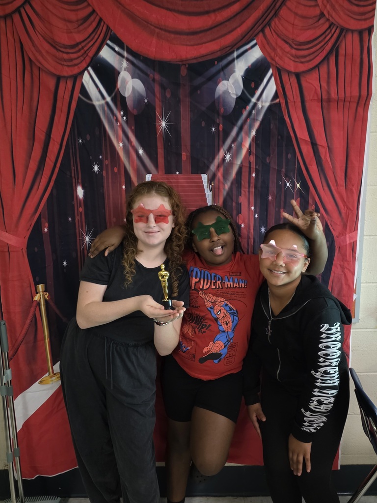 Three sixth-grade students wearing colorful star-shaped glasses pose playfully together, with one holding a small gold trophy, in front of a red-carpet backdrop.