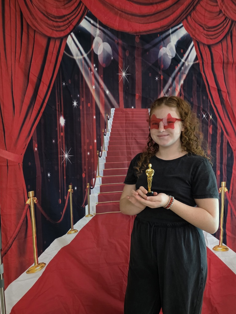 A sixth-grade student wearing red star-shaped glasses smiles while holding a small gold trophy in front of a red-carpet backdrop.