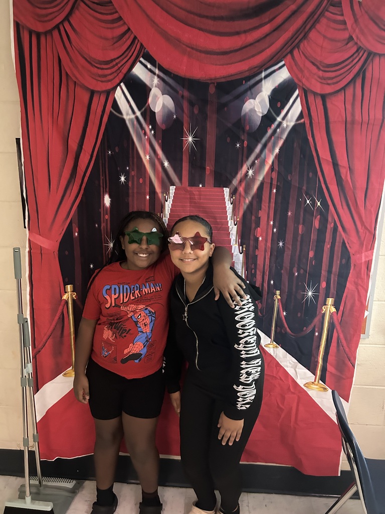 Two sixth-grade students wearing colorful star-shaped glasses smile with their arms around each other in front of a red-carpet backdrop.