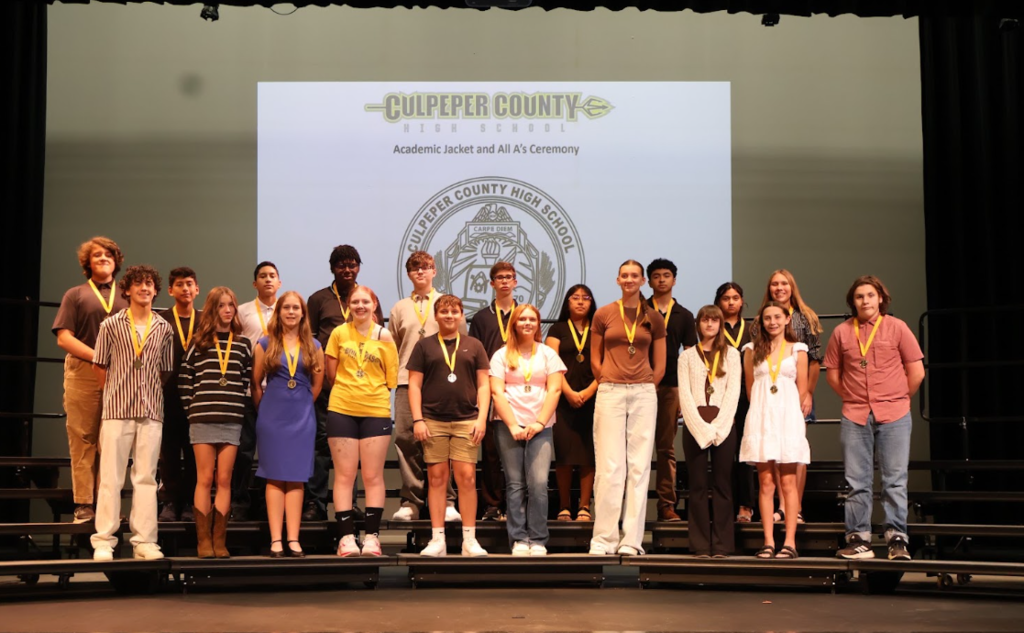 Students stand on a stage in front of a presentation screen, each wearing a medal.