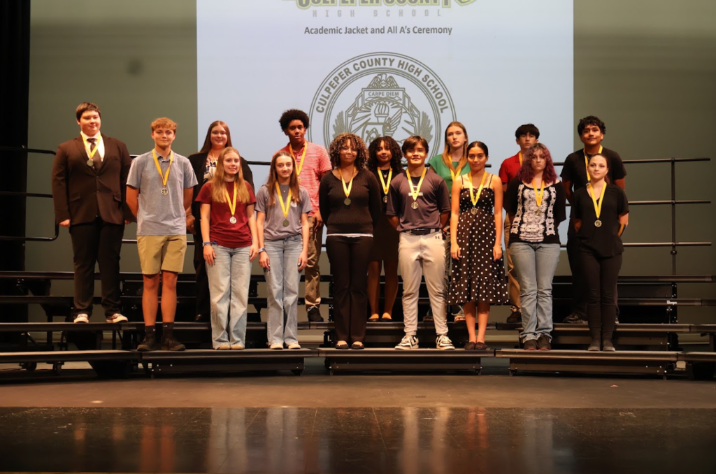 A group of students, all wearing medals, stands on a stage. A school logo is visible on a screen behind them.