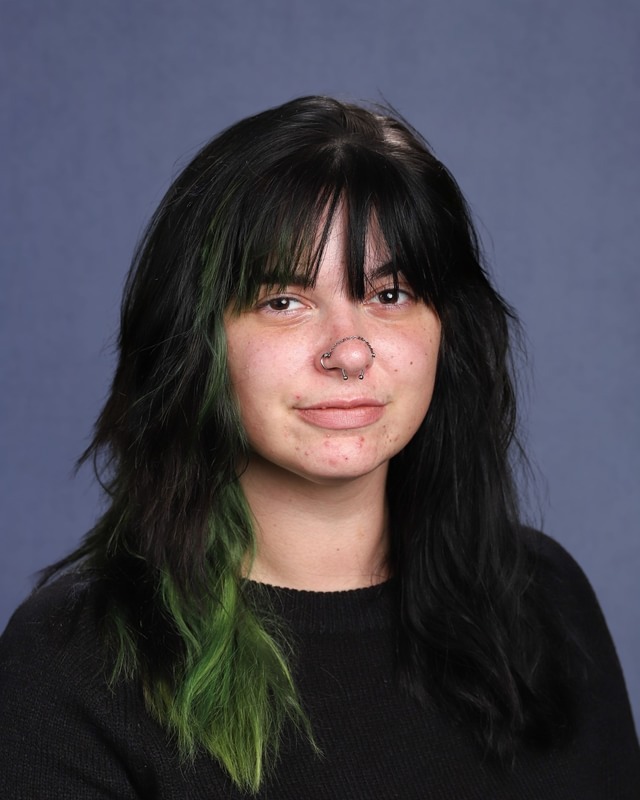 Person with black hair and green streaks, wearing a black top, standing against a blue background.
