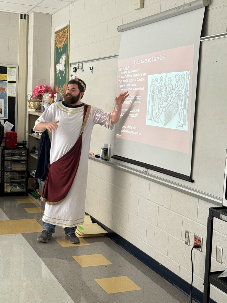 A person in a white robe and sandals presents at a whiteboard with a projector screen.