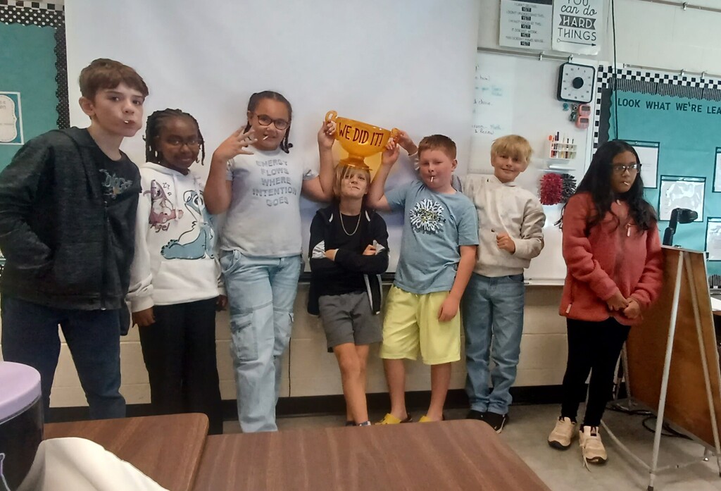 Several children, some holding yellow objects, pose for a photo in a classroom. A whiteboard and bulletin board are in the background.