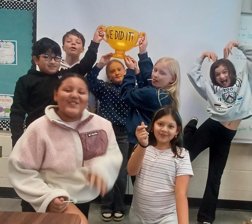 Children in a classroom hold up a large yellow trophy with "WE DID IT" written on it.