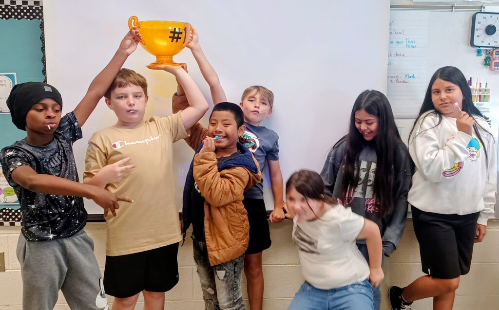 A group of young children stand in a classroom, some holding a trophy. They are smiling and appear to be posing for a photo.