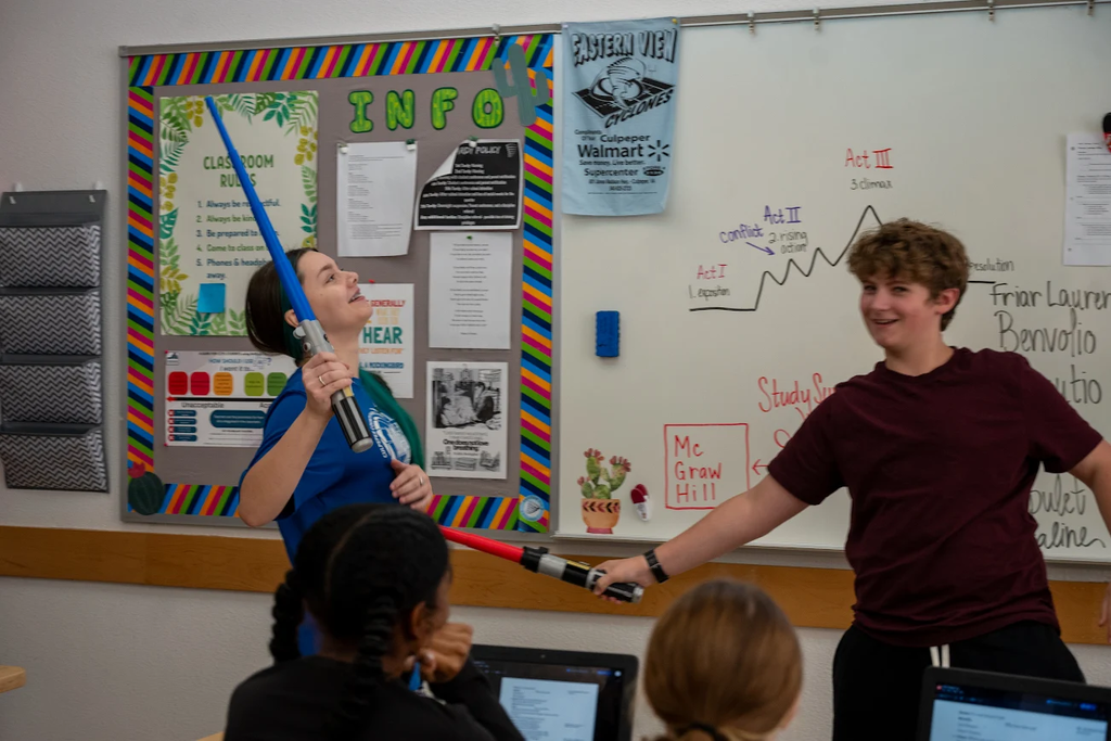 Three students in a classroom, two with toy lightsabers, one girl holding a blue blade, and boy a red one.