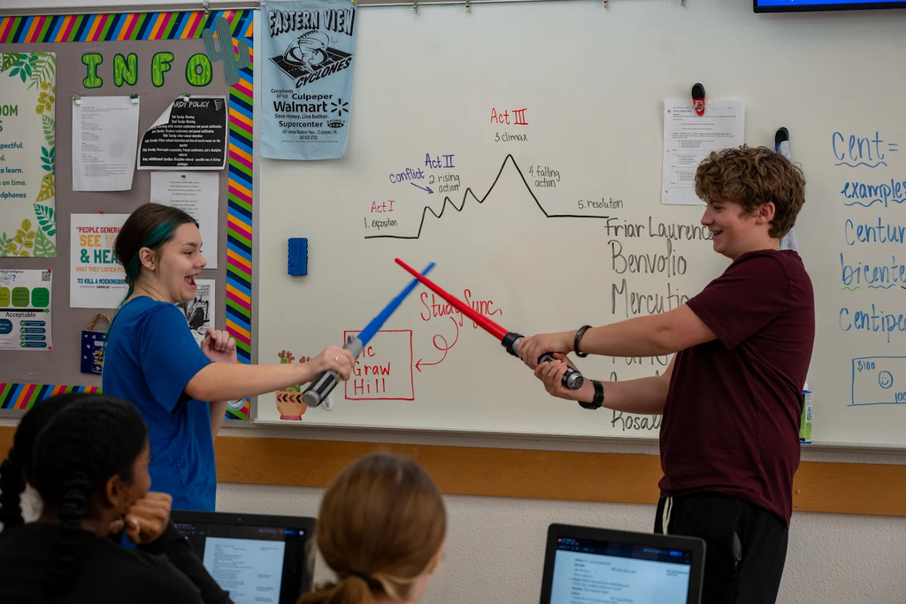 Two students with red swords reenacting a fight in a classroom with others watching. A whiteboard behind them has drawings and text.