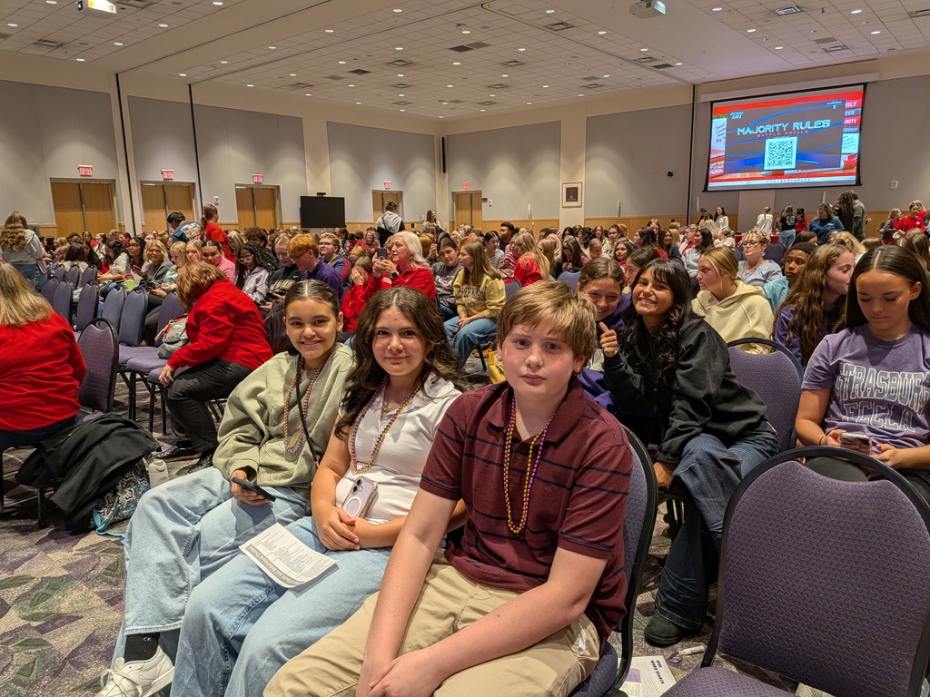 Three students sit together in a crowded auditorium with other students and adults gathered for an event.