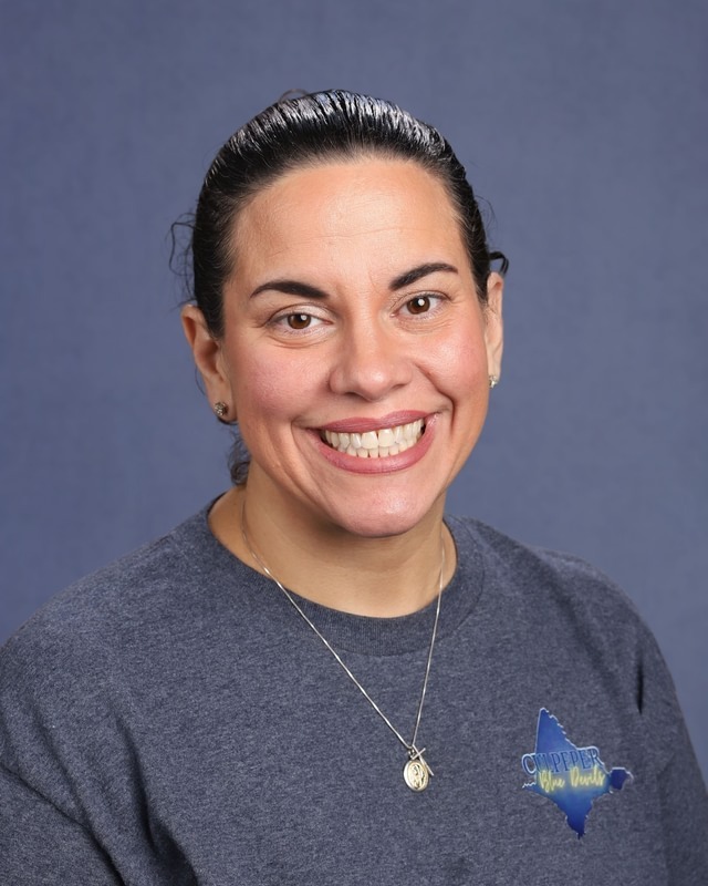 Woman in a gray shirt and necklace with a badge smiles at the camera. Light blue background.