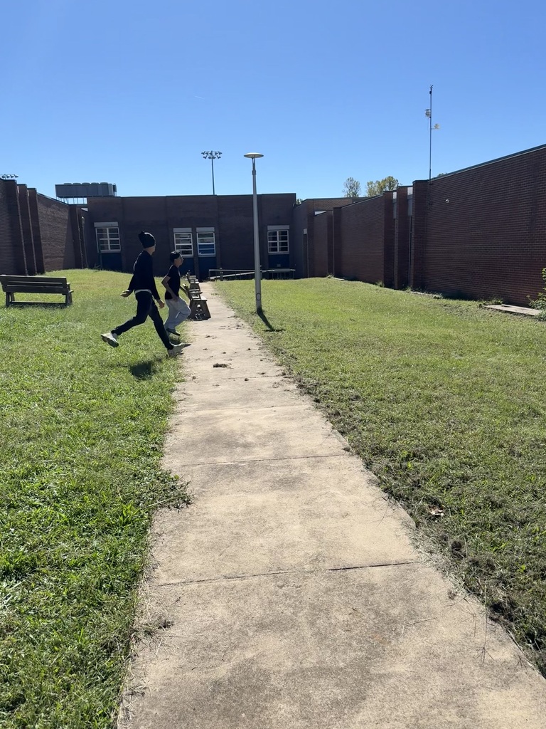 Two people run on a concrete path through a grassy area. Behind them is a building with brick walls.