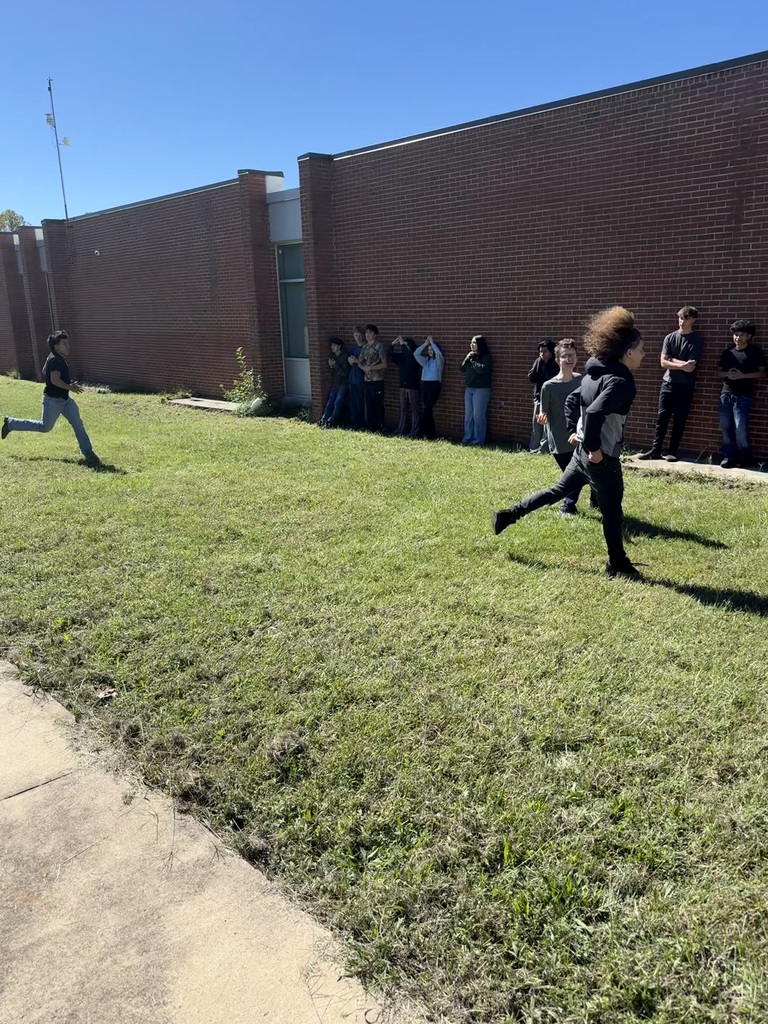 A group of people are outdoors with two running on grass. Others are standing and watching. A brick building is in the background.
