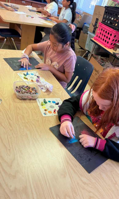 Three young girls seated at a wooden table, engaged in an art activity. One girl holds a crayon, while others use paint.