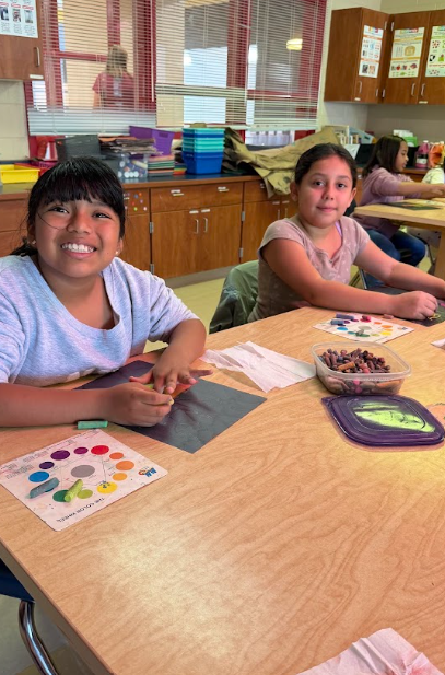 Two girls sit at a table, smiling, with art supplies. Behind them, wooden cabinets and chairs. A third girl sits at a table in the background.