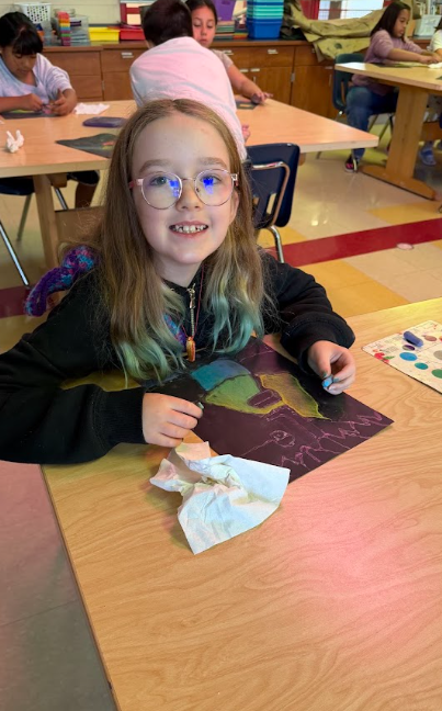 A child with glasses sits at a table, smiling while working on a colorful art project.