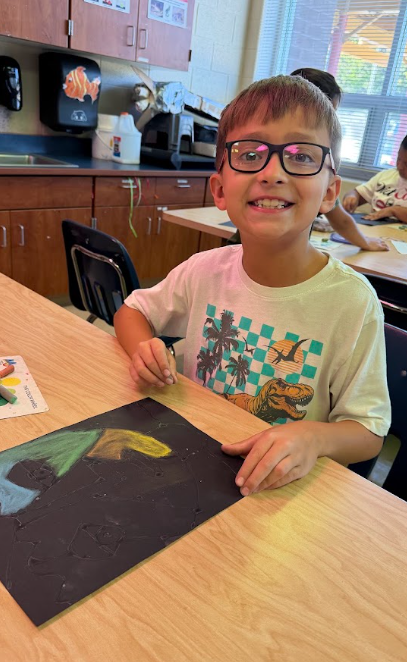 A boy with glasses sits at a desk and holds a colorful paper. Other students work behind him.