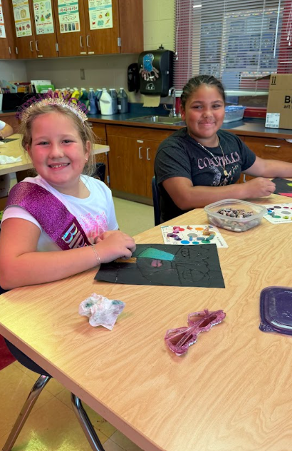 Two girls in a classroom, seated at a table with crafts, smiling and surrounded by art supplies.