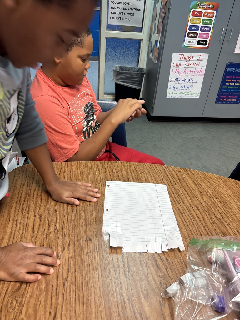 Two students working together at a table with papers, scissors, and other supplies on it.