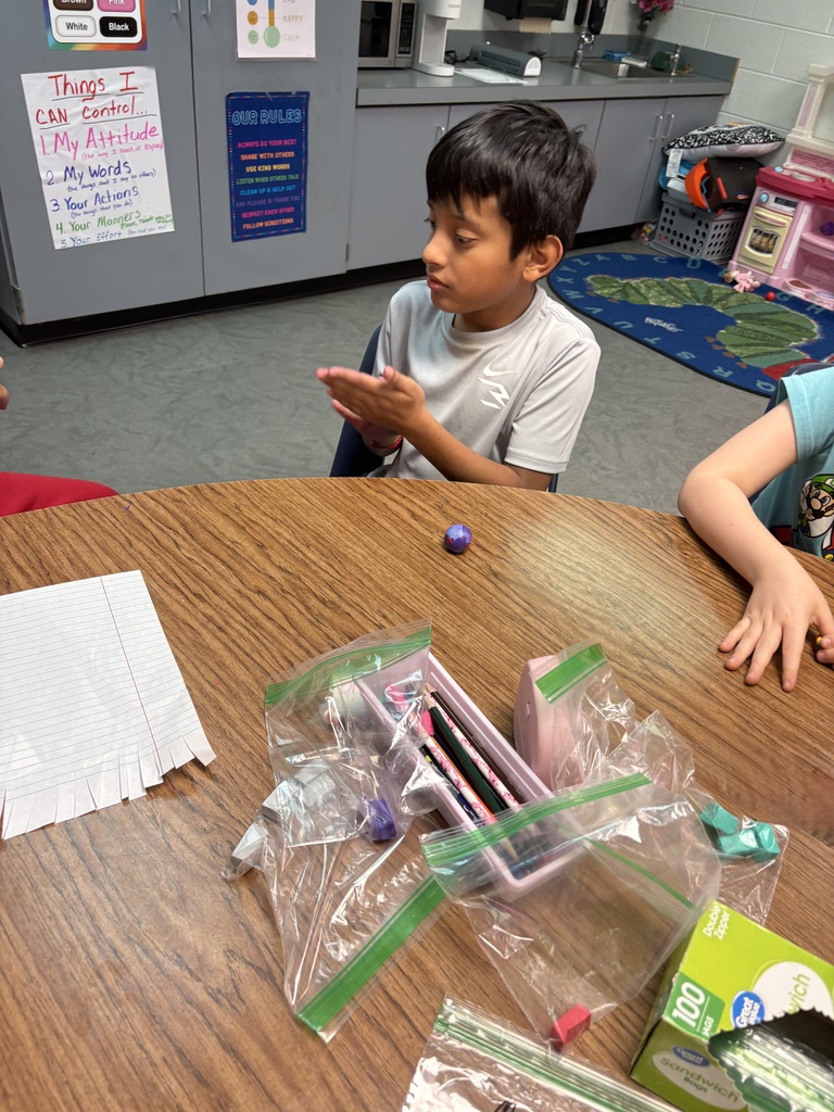 A young boy sits at a table with plastic bags and other objects, looking down. A second child sits beside him.