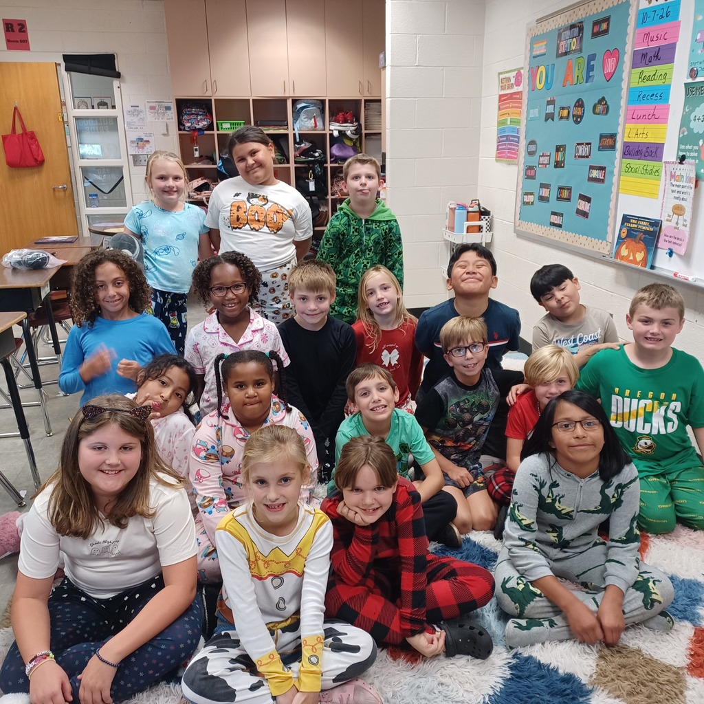 A classroom filled with children in pajamas, seated or standing. Some are smiling, and the floor is covered with a white carpet.