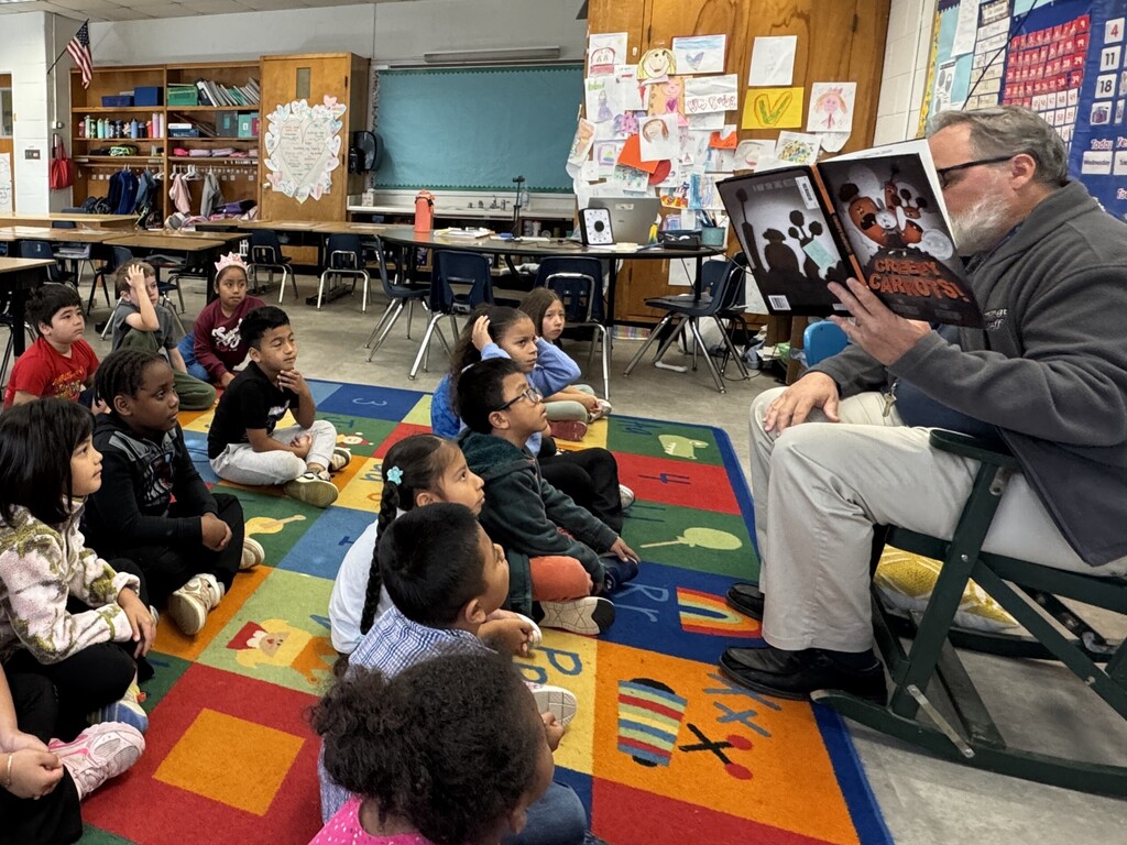Man seated in chair reads book to attentive children sitting on rug in classroom with tables and chairs.