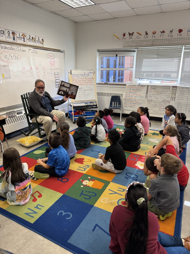 An adult sits on a chair and reads a book to children on a colorful floor mat.