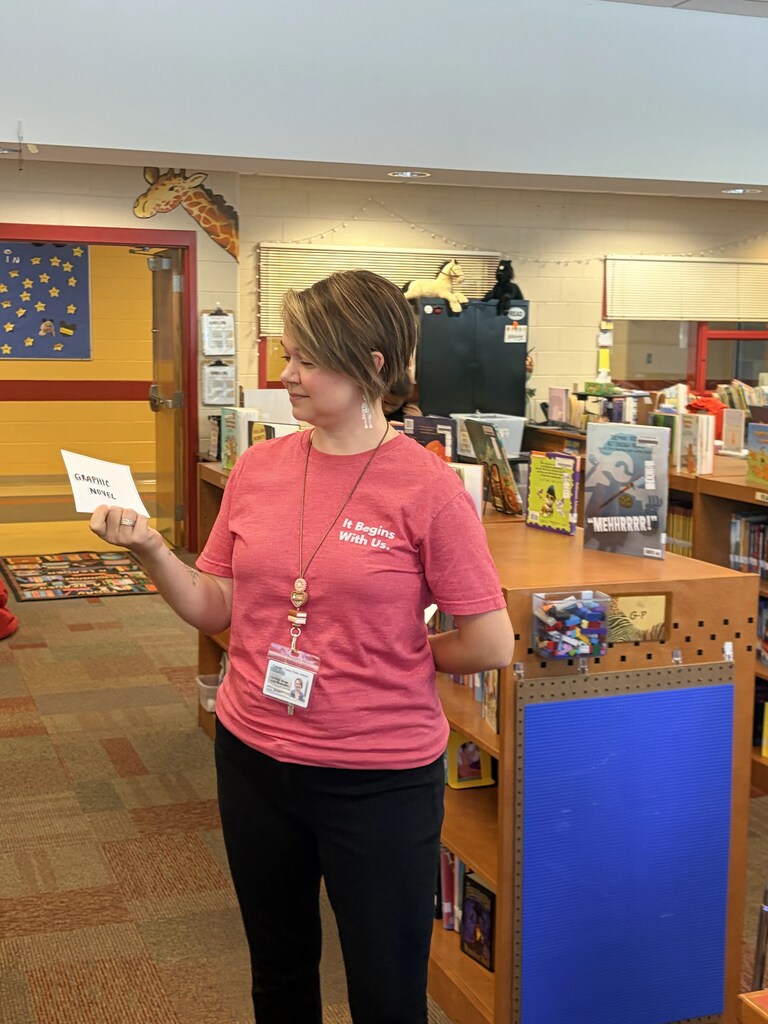 A woman in a pink shirt holds a card in a library with shelves and displays.