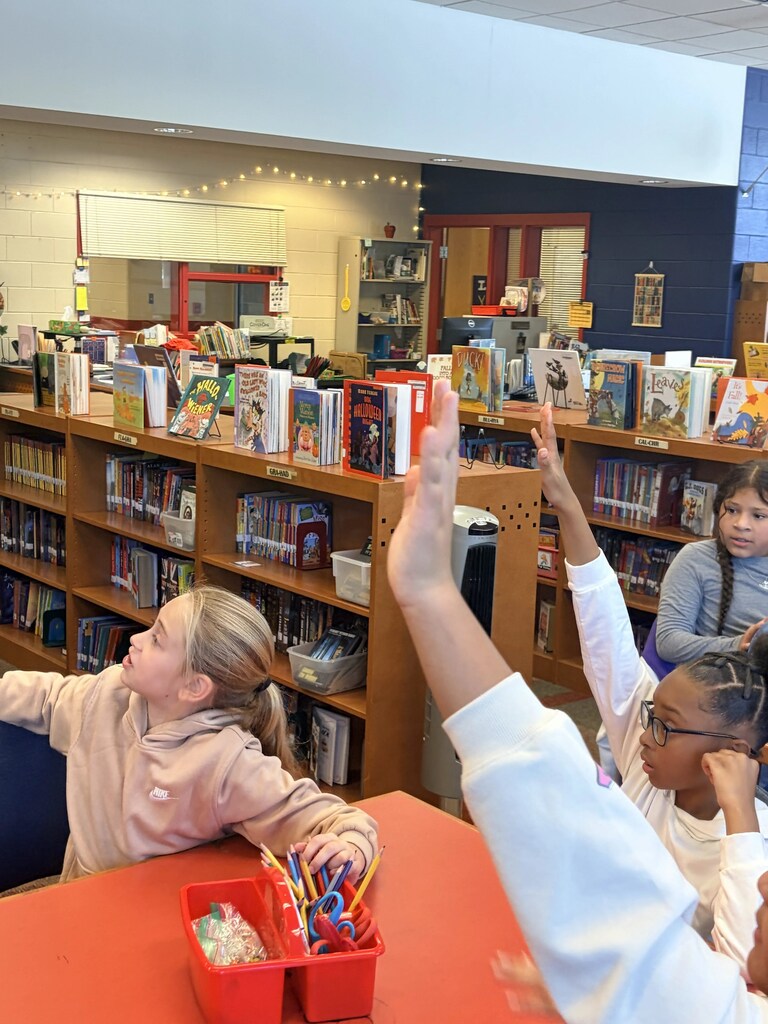 Four children sitting at a table in a library. Two children raise their hands. Books fill shelves in the background.
