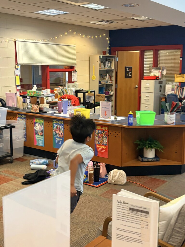 Child near a desk in a library, holding a whiteboard. Behind is a counter with books and items.