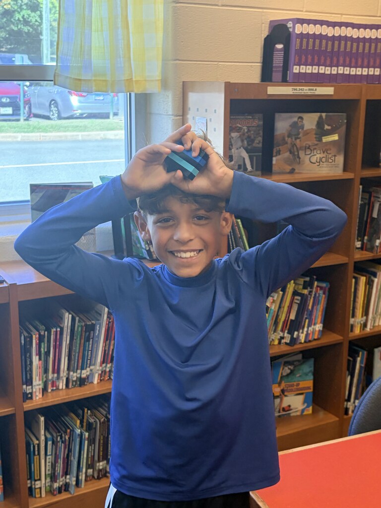 A boy in a blue long-sleeve shirt poses with a handheld device near a bookshelf.