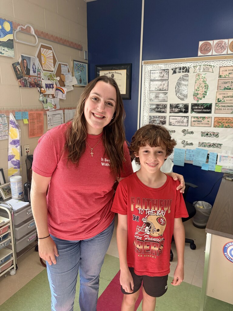 A woman and a boy stand in a classroom. She wears a red shirt, he wears a red shirt with text. Behind them is a bulletin board.