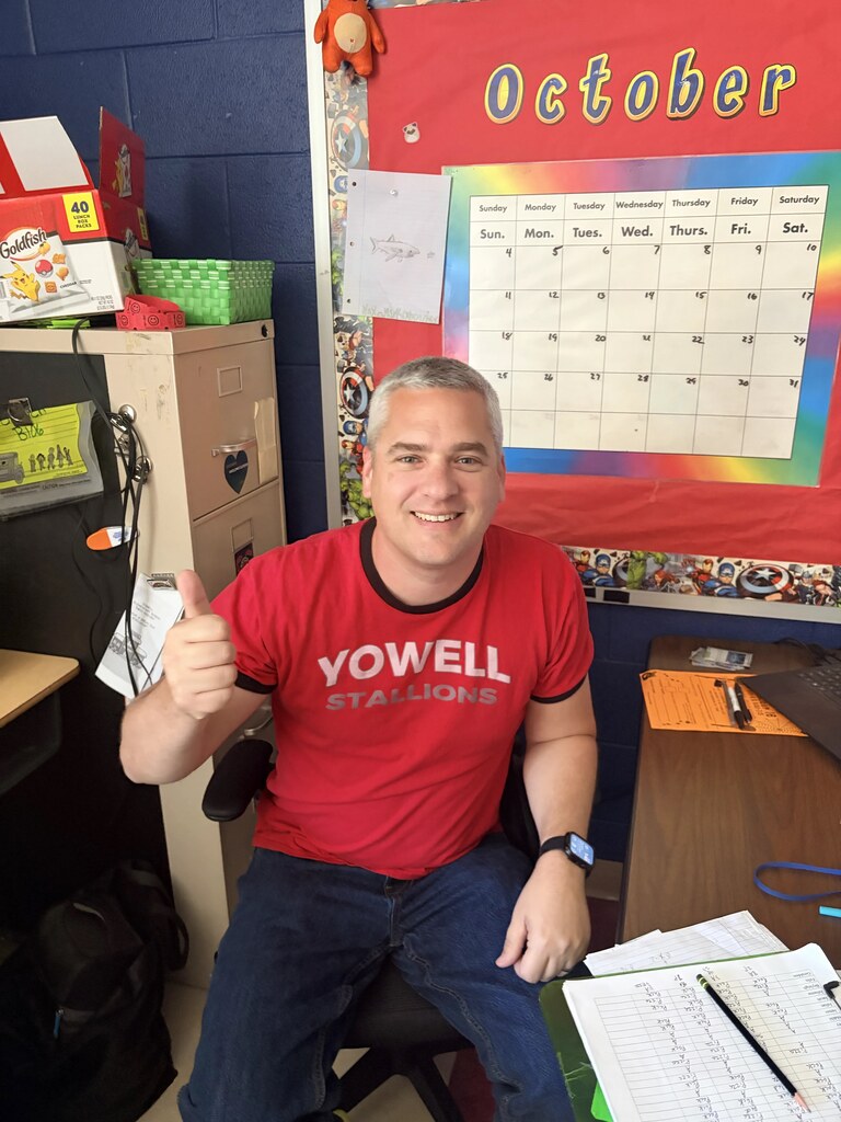 A man sits in an office chair, giving a thumbs up. He wears a red t-shirt with white letters.