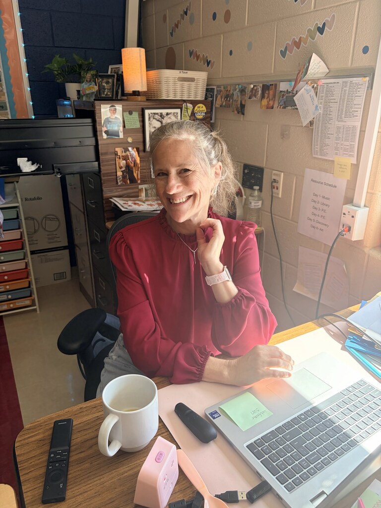 A person sits at a desk with a laptop and coffee mug, smiling, in a room with shelves, wall art, and an open door.