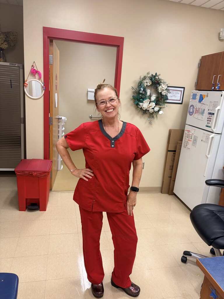 A woman in red scrubs stands in a room with a refrigerator, desk, and decorative wreath.