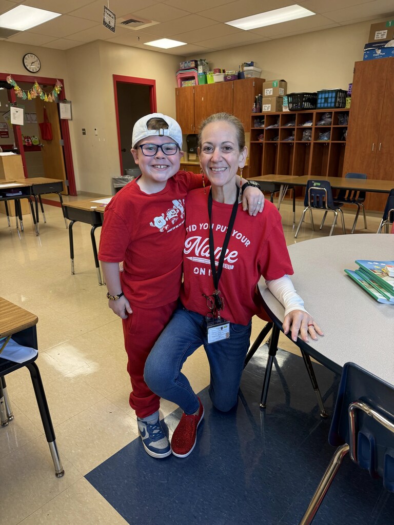 An adult and a child wear red t-shirts. They pose in a classroom. The floor is covered with carpet.