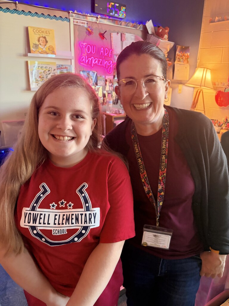 A girl with blonde hair in a red shirt stands beside an adult woman with glasses.