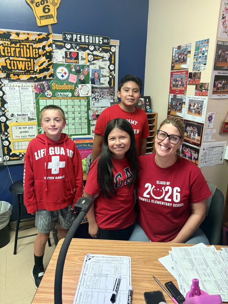 Four people in red shirts, standing and sitting in a classroom with a table and bulletin board.
